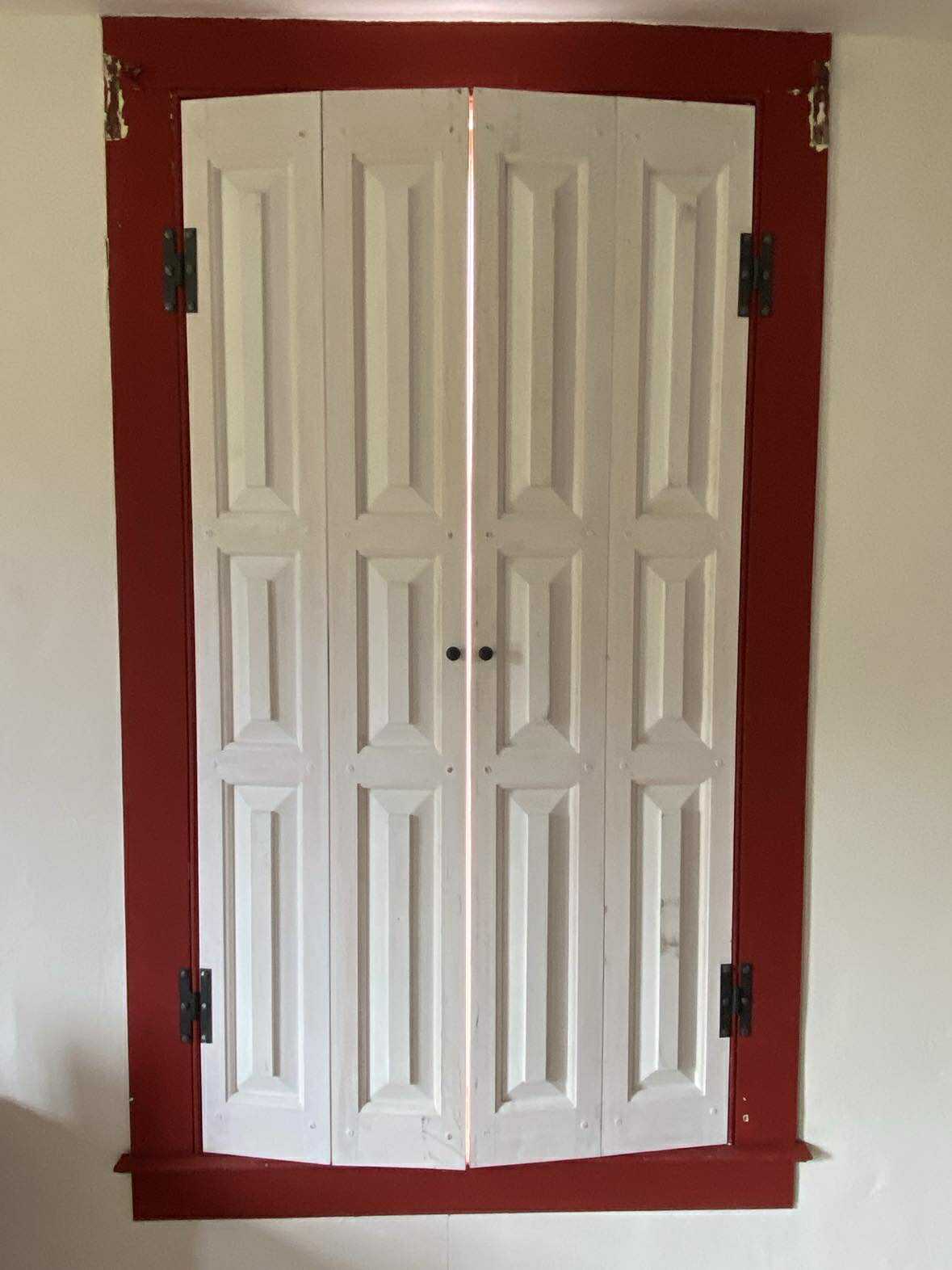 Two white raised-panel shutters installed in bold red window trim with wall lantern