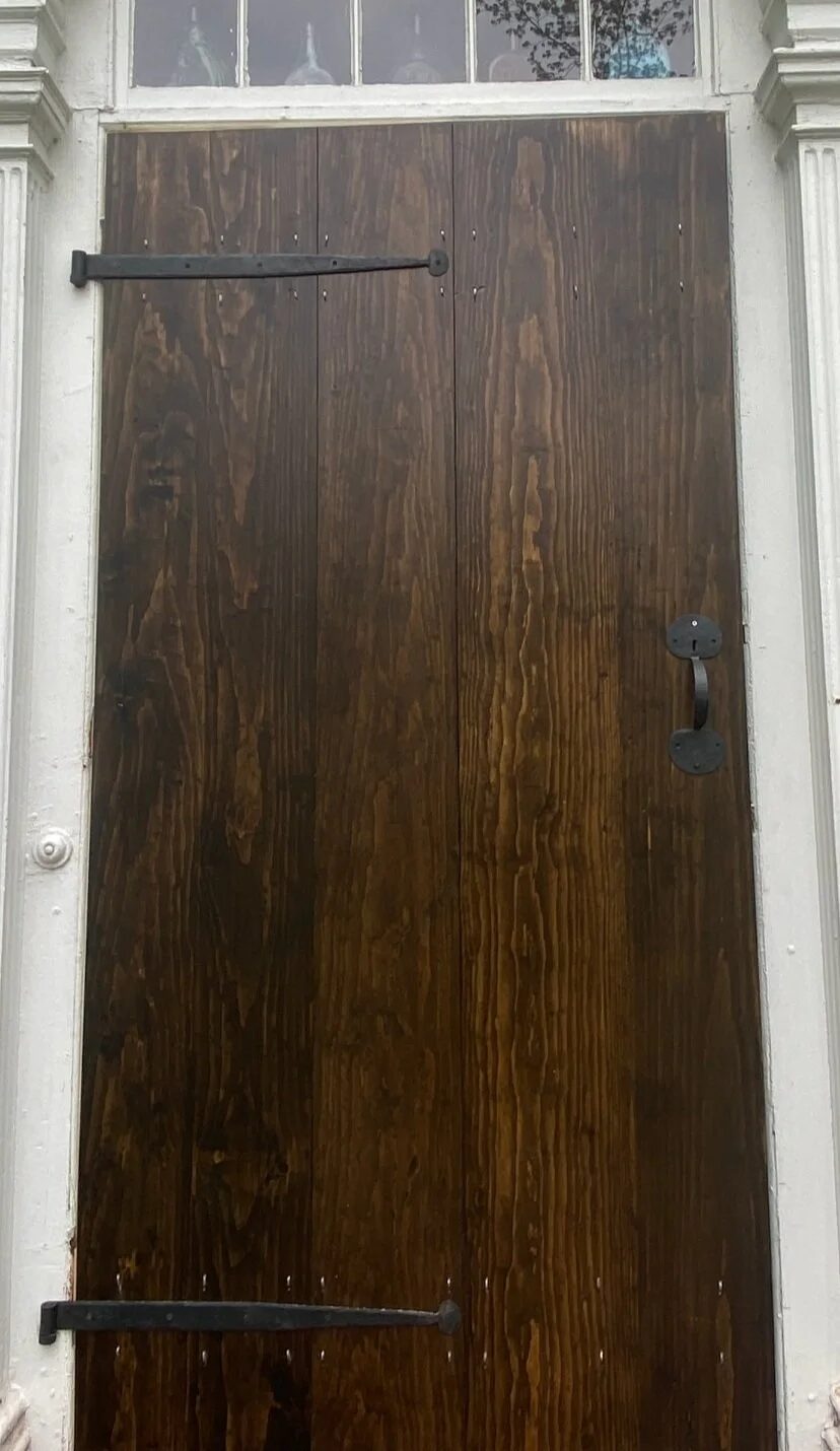 Dark stained board-and-batten storm door close-up showing black strap hinges and period iron latch